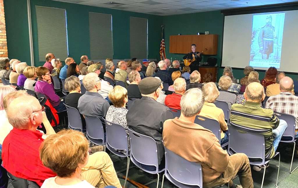 Crowd sitting in a room, man with guitar at front of room. Projector screen with image of a solider to right of the man.